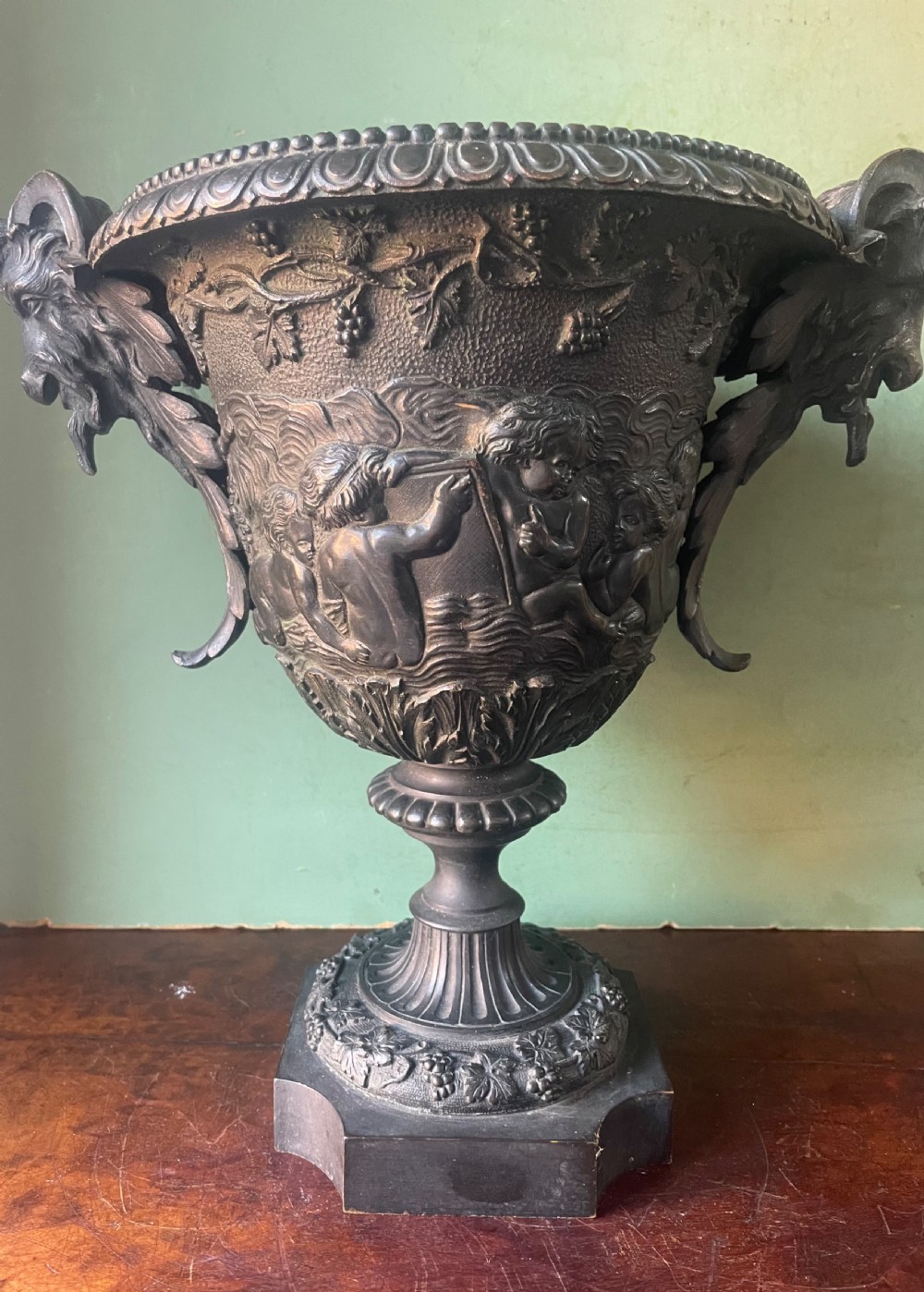 A 19th-century French dark bronze campana-shaped urn featuring relief carving of putti, satyr mask handles, and a square pedestal base. View XVIII