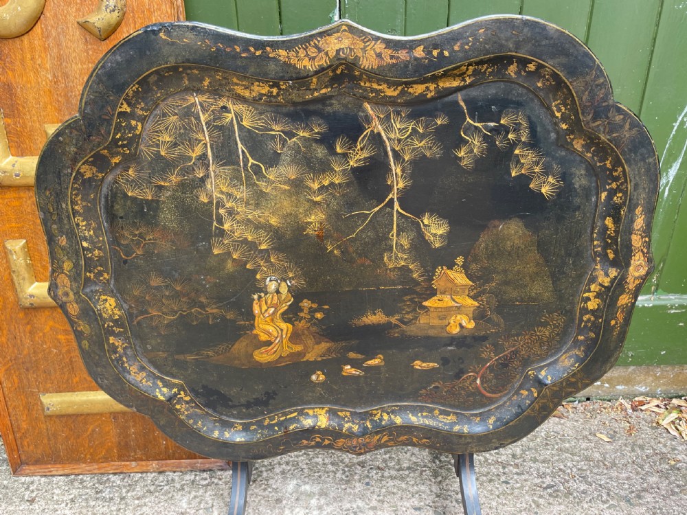 A 19th-century black lacquered papier-mache tray table featuring gilded Chinoiserie landscape decoration and scalloped edges.