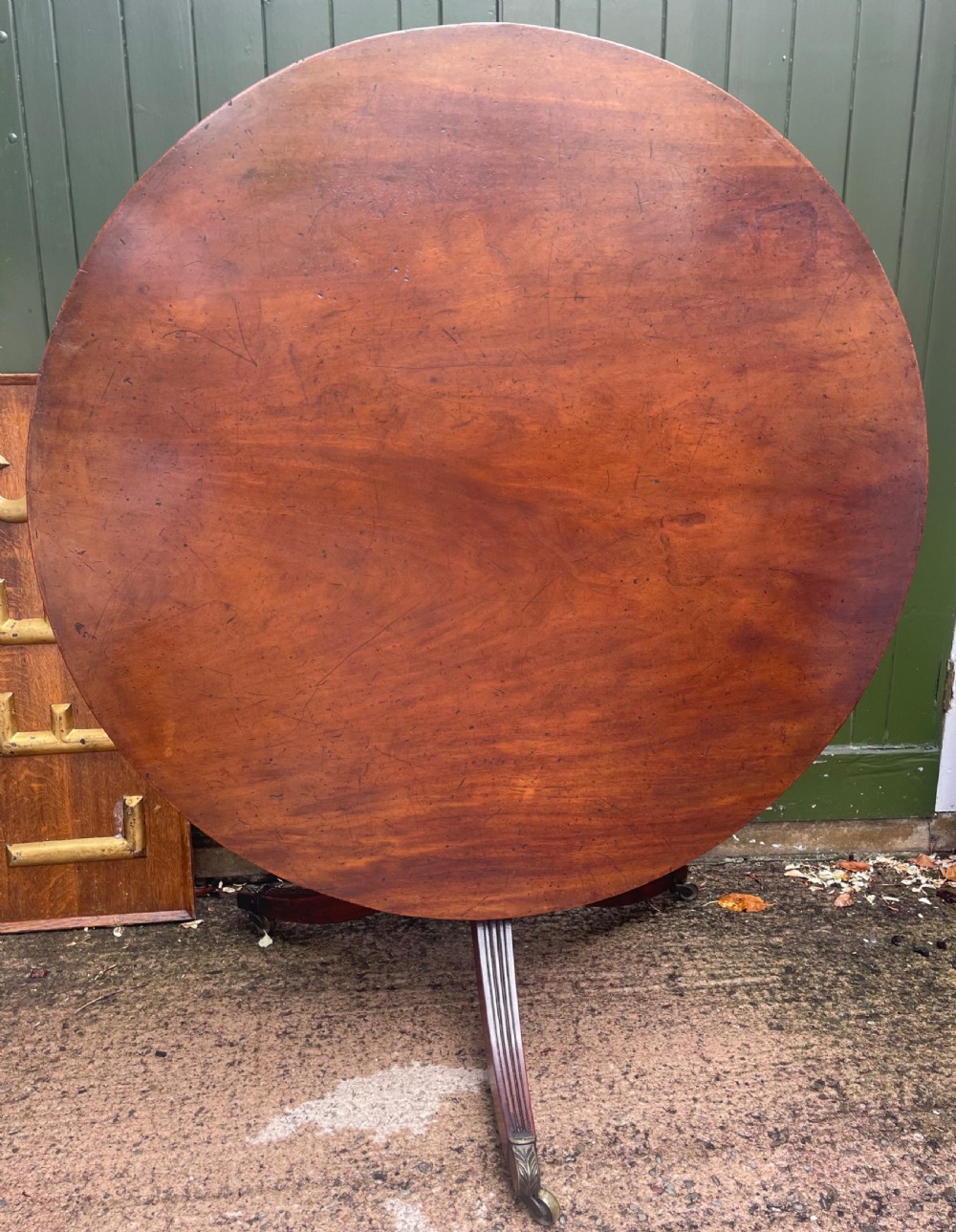A late George III period circular mahogany tilt-top table shown in a vertical position, featuring a single-piece top and reeded tripod legs with brass castors. View II
