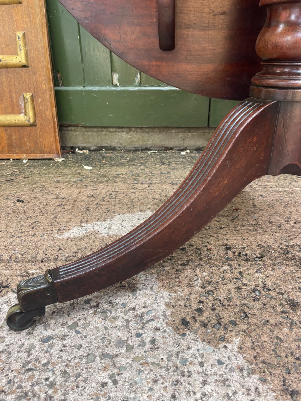 A late George III period circular mahogany tilt-top table shown in a vertical position, featuring a single-piece top and reeded tripod legs with brass castors. View VIII