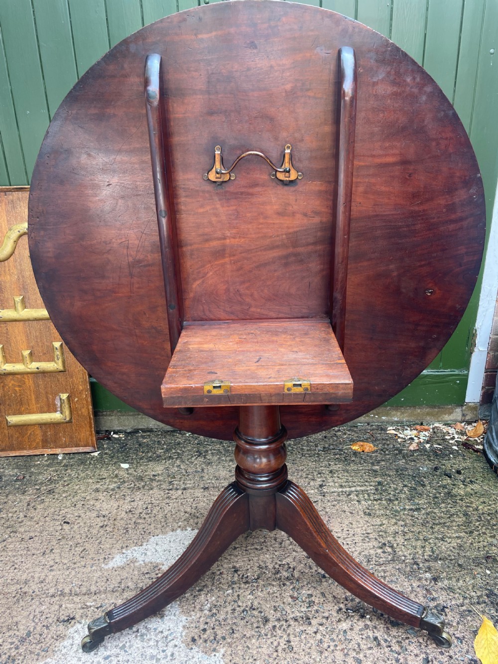 A late George III period circular mahogany tilt-top table shown in a vertical position, featuring a single-piece top and reeded tripod legs with brass castors. View X