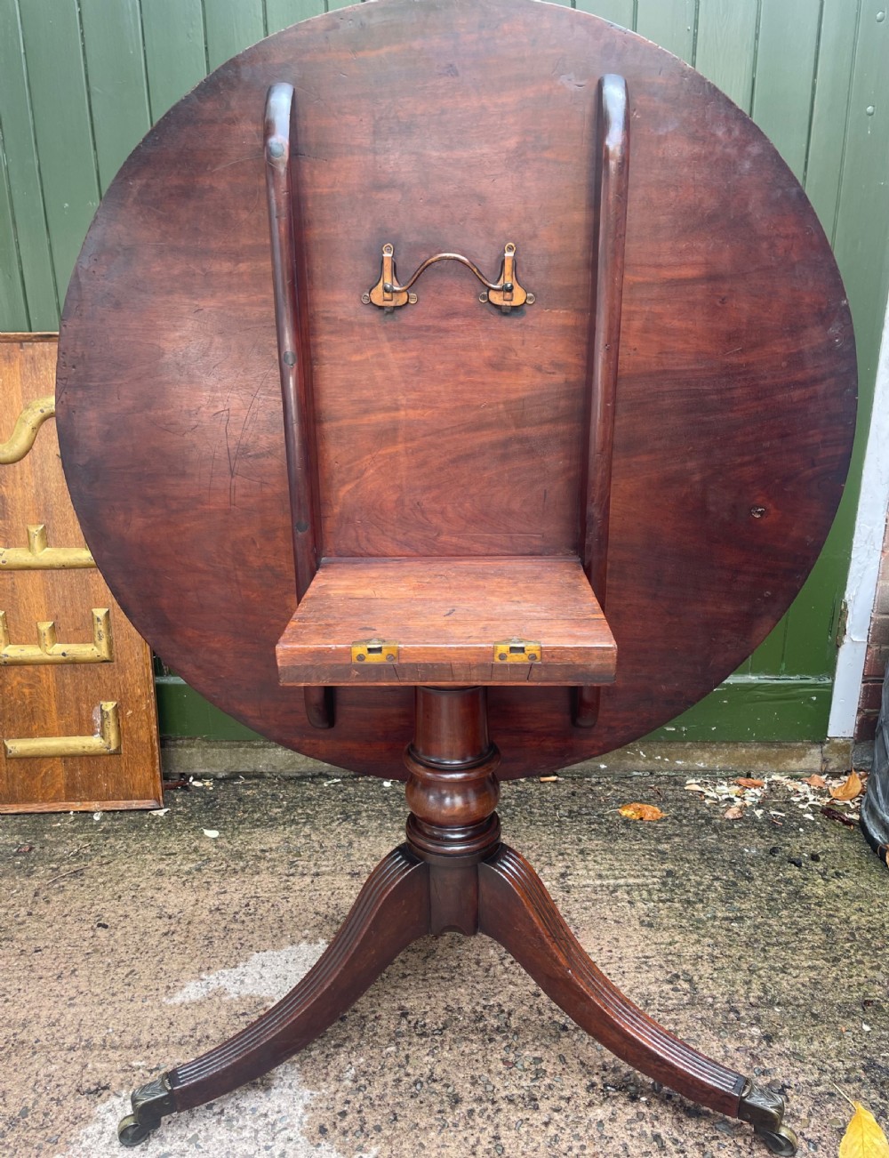 A late George III period circular mahogany tilt-top table shown in a vertical position, featuring a single-piece top and reeded tripod legs with brass castors. View XII