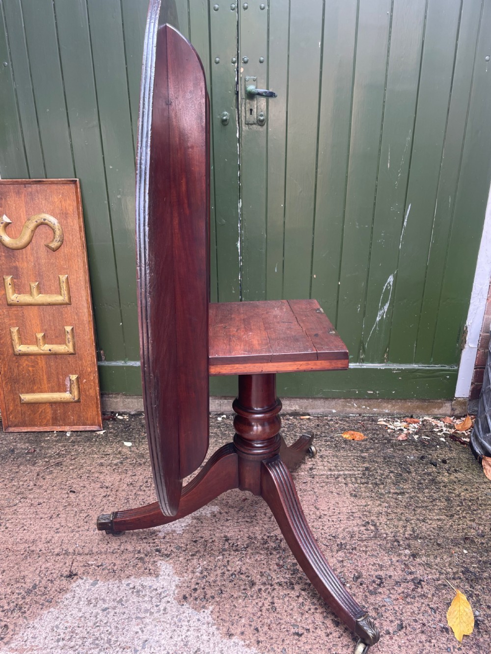 A late George III period circular mahogany tilt-top table shown in a vertical position, featuring a single-piece top and reeded tripod legs with brass castors. View XIV