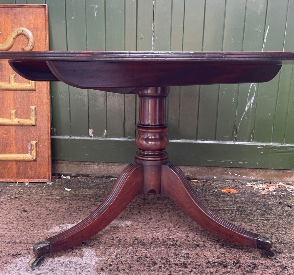 A late George III period circular mahogany tilt-top table shown in a vertical position, featuring a single-piece top and reeded tripod legs with brass castors. View XV