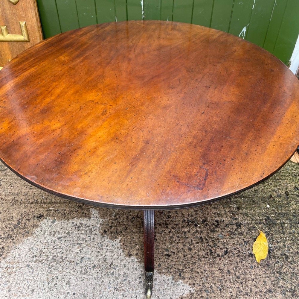 A late George III period circular mahogany tilt-top table shown in a vertical position, featuring a single-piece top and reeded tripod legs with brass castors. View XVII