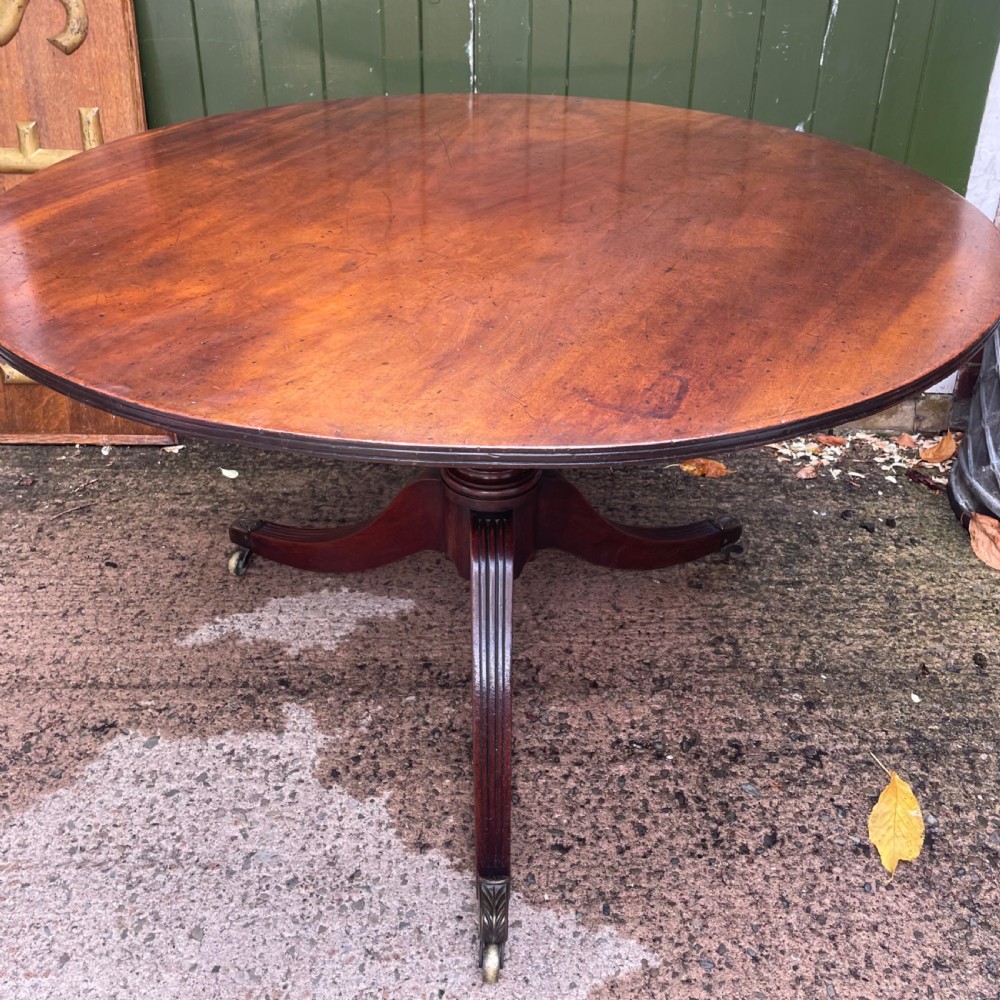 A late George III period circular mahogany tilt-top table shown in a vertical position, featuring a single-piece top and reeded tripod legs with brass castors. View XIX