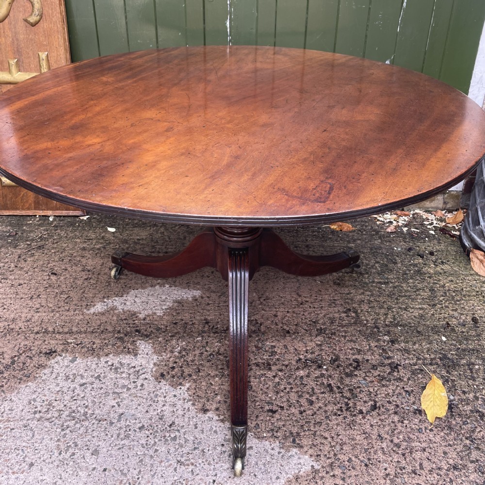 A late George III period circular mahogany tilt-top table shown in a vertical position, featuring a single-piece top and reeded tripod legs with brass castors. View III