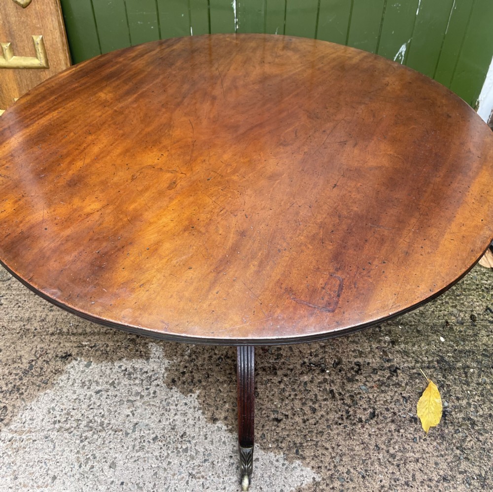 A late George III period circular mahogany tilt-top table shown in a vertical position, featuring a single-piece top and reeded tripod legs with brass castors. View XXIII