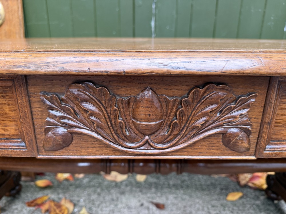 An early 19th-century William IV oak library table featuring a central carved acorn frieze panel, scrolled trestle supports, and a turned stretcher base. View II