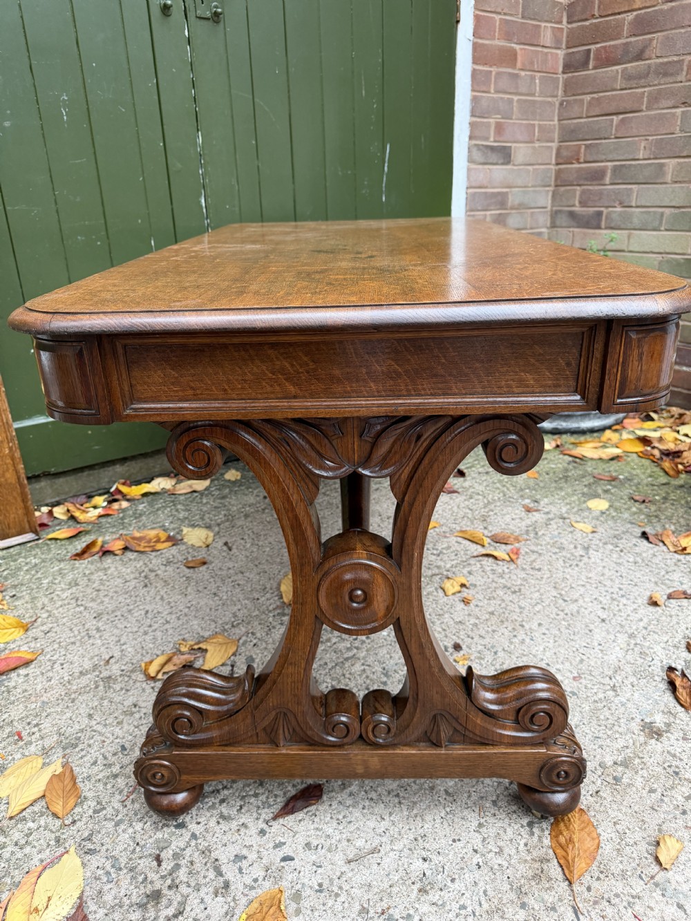 An early 19th-century William IV oak library table featuring a central carved acorn frieze panel, scrolled trestle supports, and a turned stretcher base. View V