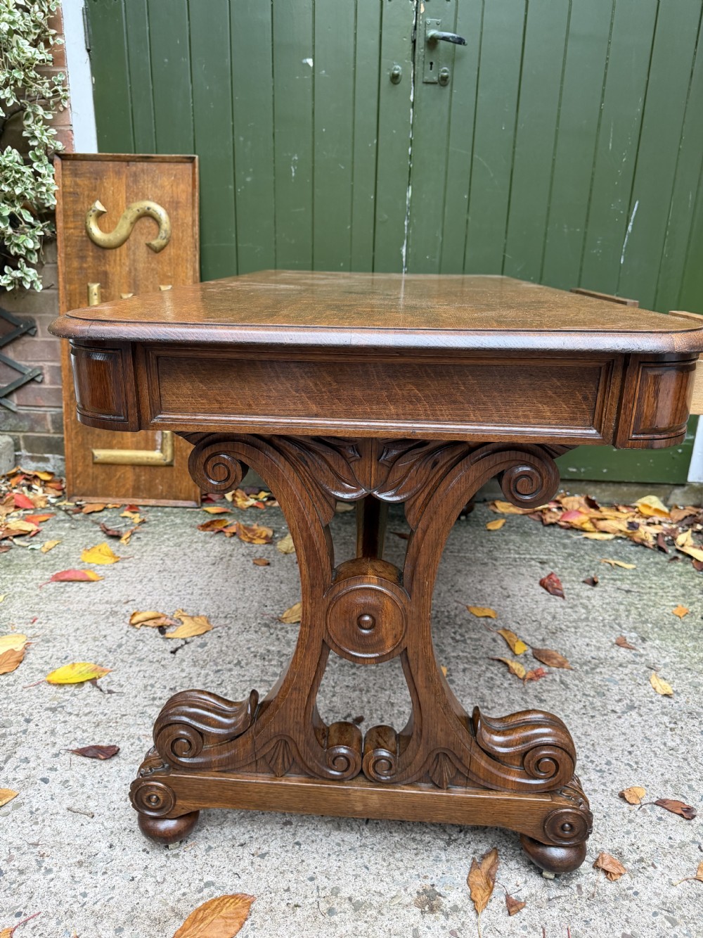 An early 19th-century William IV oak library table featuring a central carved acorn frieze panel, scrolled trestle supports, and a turned stretcher base. View VIII