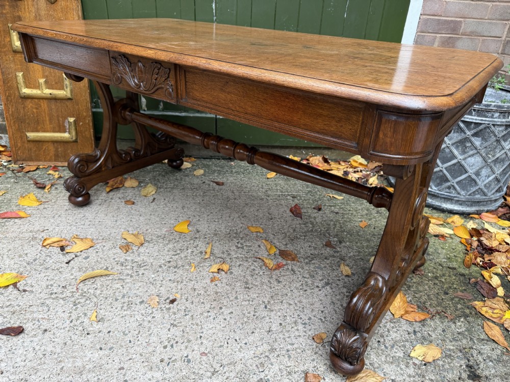 An early 19th-century William IV oak library table featuring a central carved acorn frieze panel, scrolled trestle supports, and a turned stretcher base. View XV