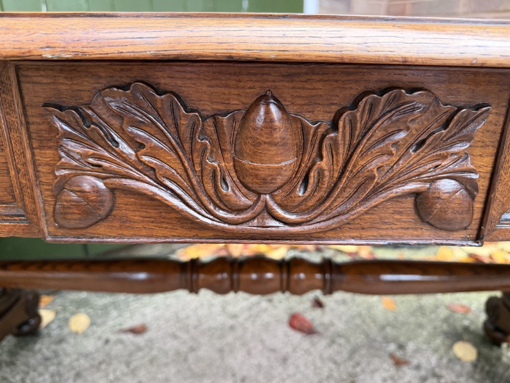 An early 19th-century William IV oak library table featuring a central carved acorn frieze panel, scrolled trestle supports, and a turned stretcher base. View XVI