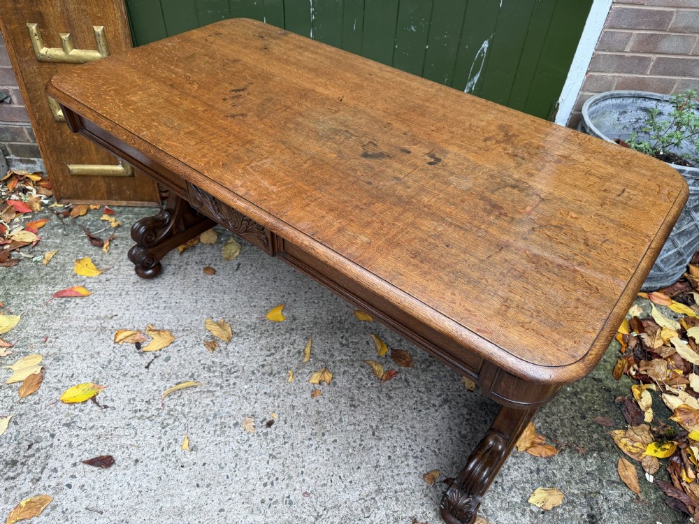 An early 19th-century William IV oak library table featuring a central carved acorn frieze panel, scrolled trestle supports, and a turned stretcher base. View XX