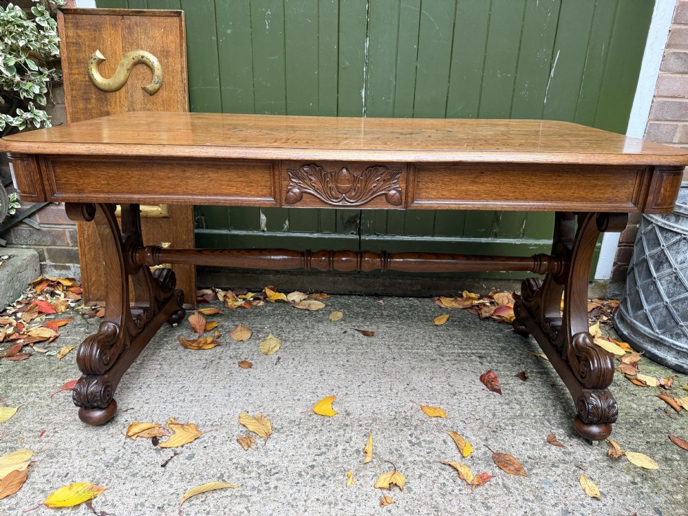 An early 19th-century William IV oak library table featuring a central carved acorn frieze panel, scrolled trestle supports, and a turned stretcher base. View XXII