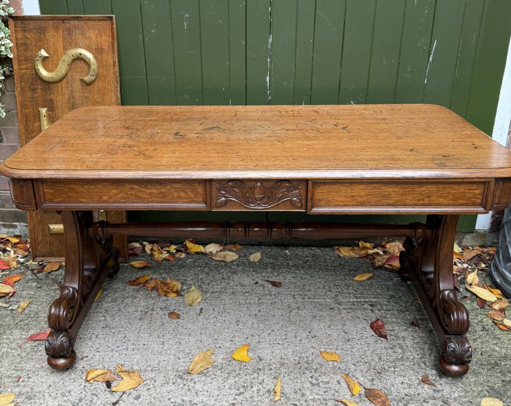 An early 19th-century William IV oak library table featuring a central carved acorn frieze panel, scrolled trestle supports, and a turned stretcher base. View XXVI
