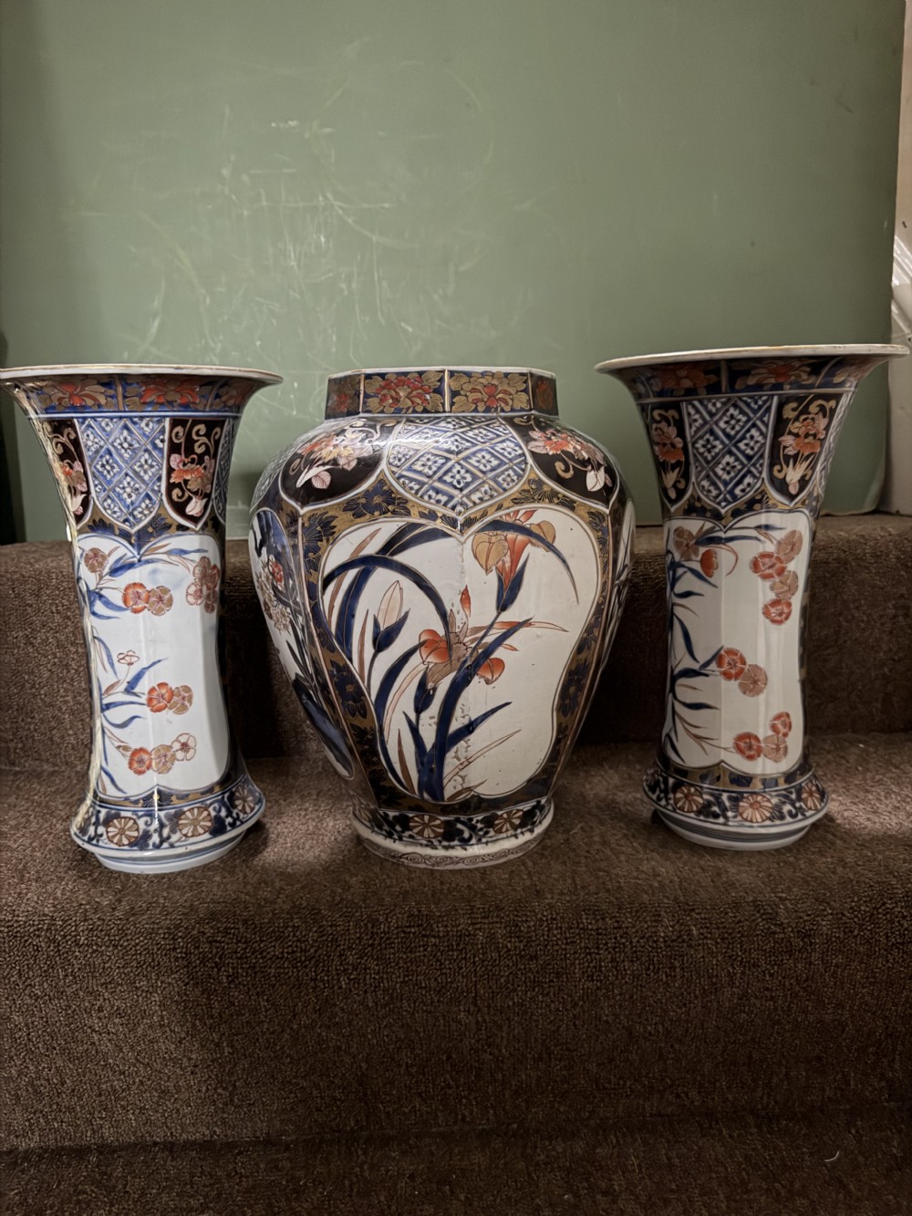 A three-piece Japanese Imari porcelain garniture featuring a central octagonal baluster jar flanked by two trumpet-shaped beaker vases, decorated with floral panels in cobalt blue, iron red, and gold. View XII