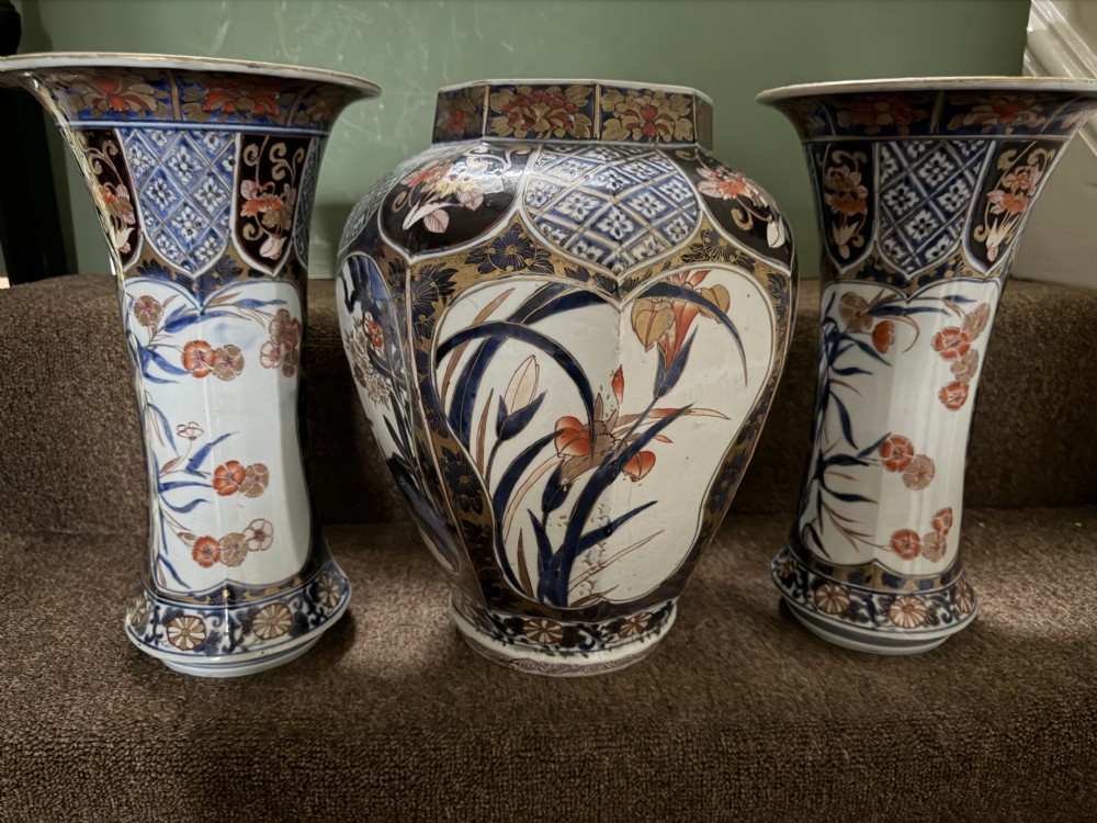 A three-piece Japanese Imari porcelain garniture featuring a central octagonal baluster jar flanked by two trumpet-shaped beaker vases, decorated with floral panels in cobalt blue, iron red, and gold. View XVI