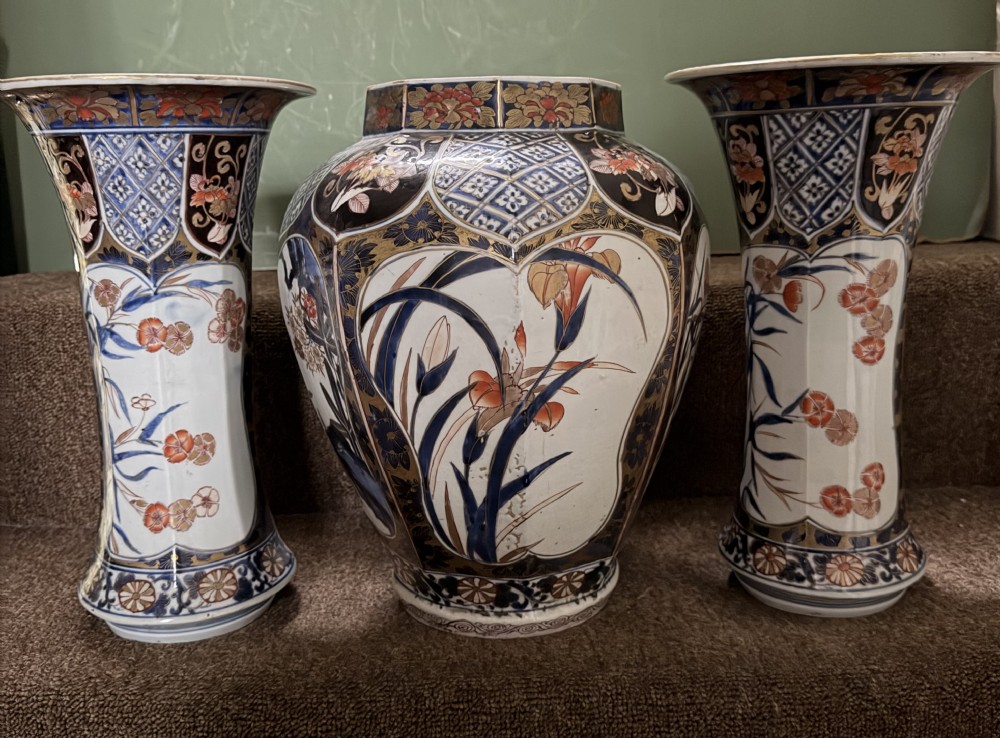 A three-piece Japanese Imari porcelain garniture featuring a central octagonal baluster jar flanked by two trumpet-shaped beaker vases, decorated with floral panels in cobalt blue, iron red, and gold. View XVII