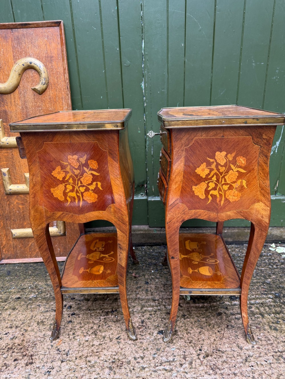 A pair of French marquetry bedside tables featuring three drawers with floral inlays, brass mounts, curved legs, and a lower tier shelf. View X