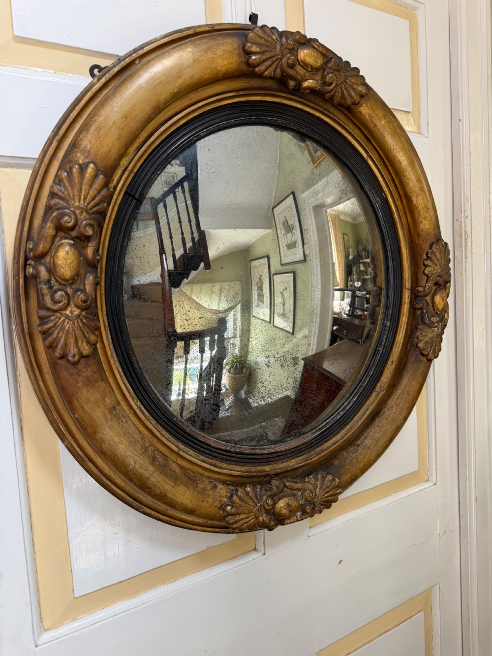 An early 19th-century Regency circular giltwood convex wall mirror featuring carved shell motifs, an ebonised reeded inner slip, and foxed mirror glass.