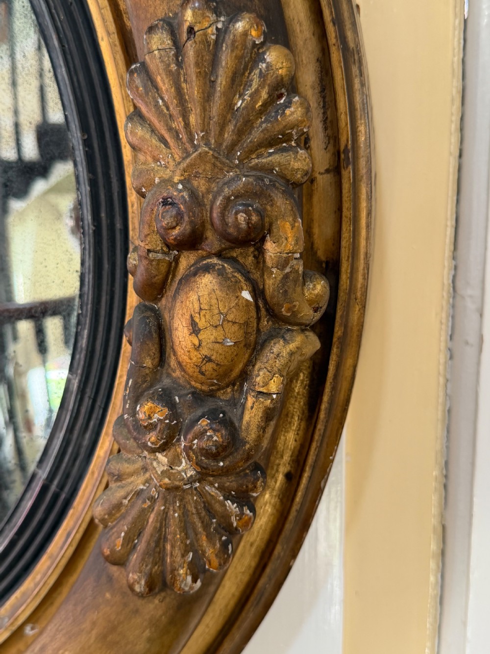 An early 19th-century Regency circular giltwood convex wall mirror featuring carved shell motifs, an ebonised reeded inner slip, and foxed mirror glass. View VI