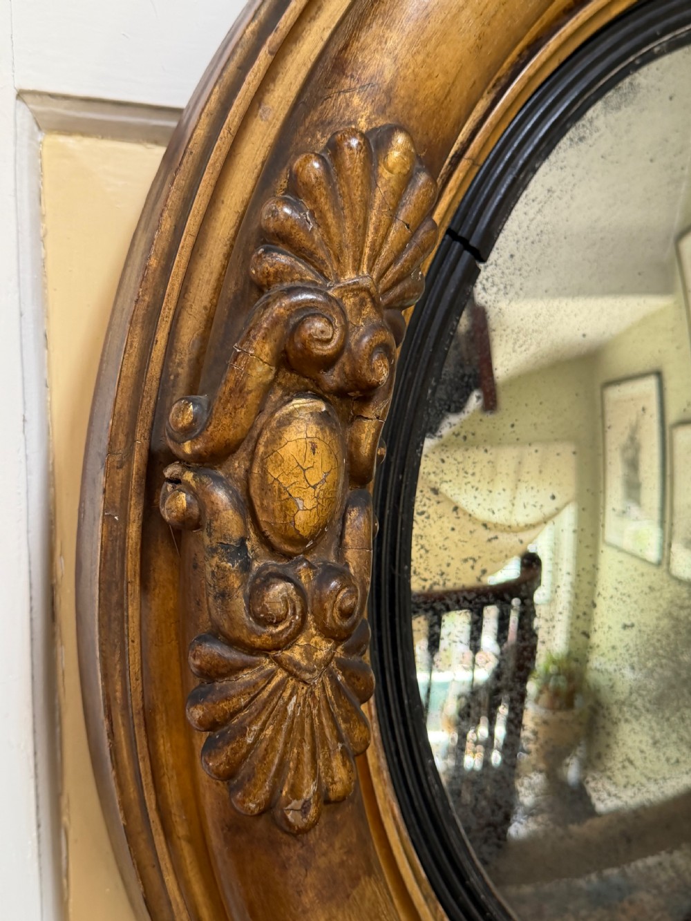 An early 19th-century Regency circular giltwood convex wall mirror featuring carved shell motifs, an ebonised reeded inner slip, and foxed mirror glass. View VII