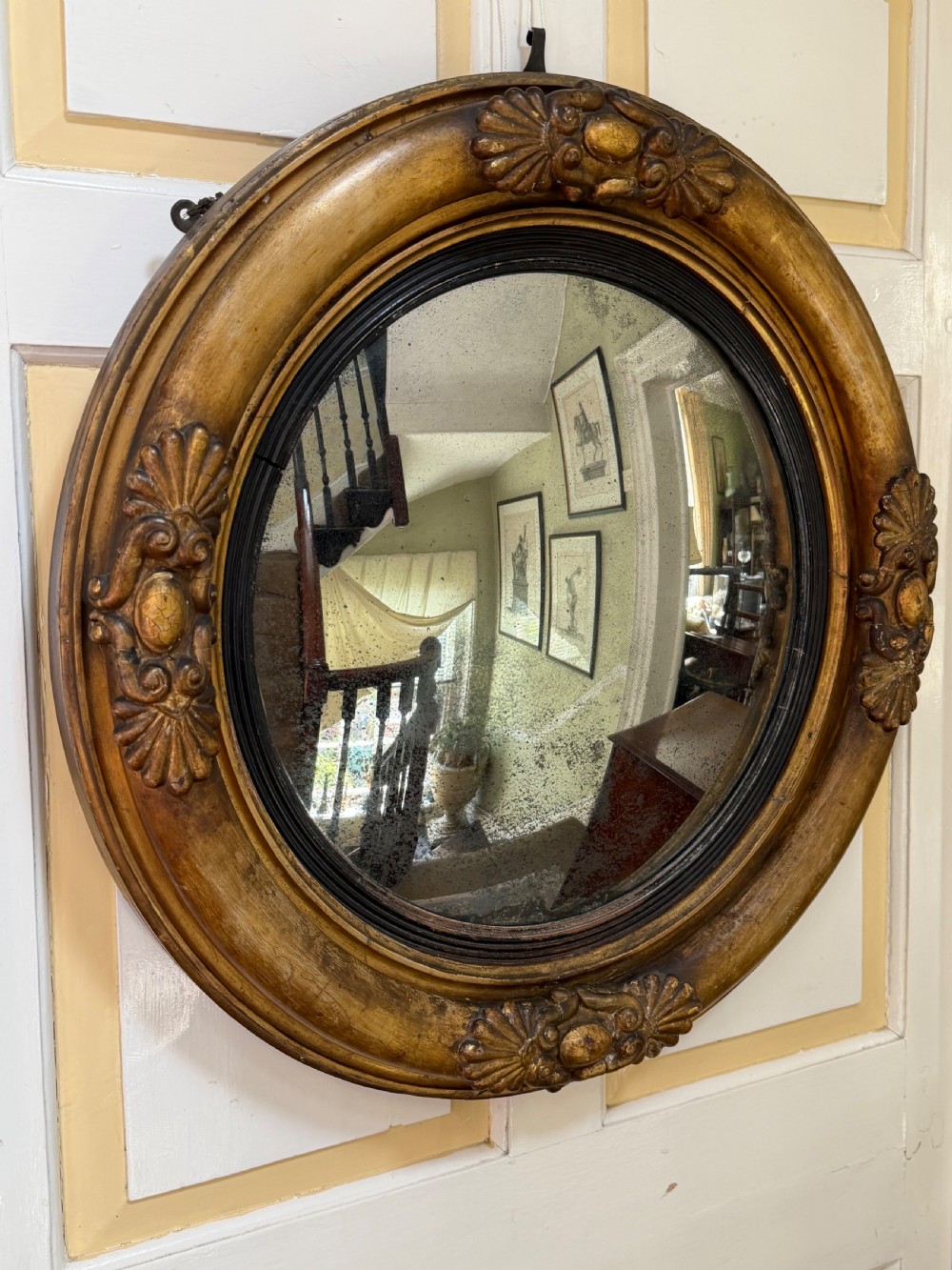 An early 19th-century Regency circular giltwood convex wall mirror featuring carved shell motifs, an ebonised reeded inner slip, and foxed mirror glass. View VIII