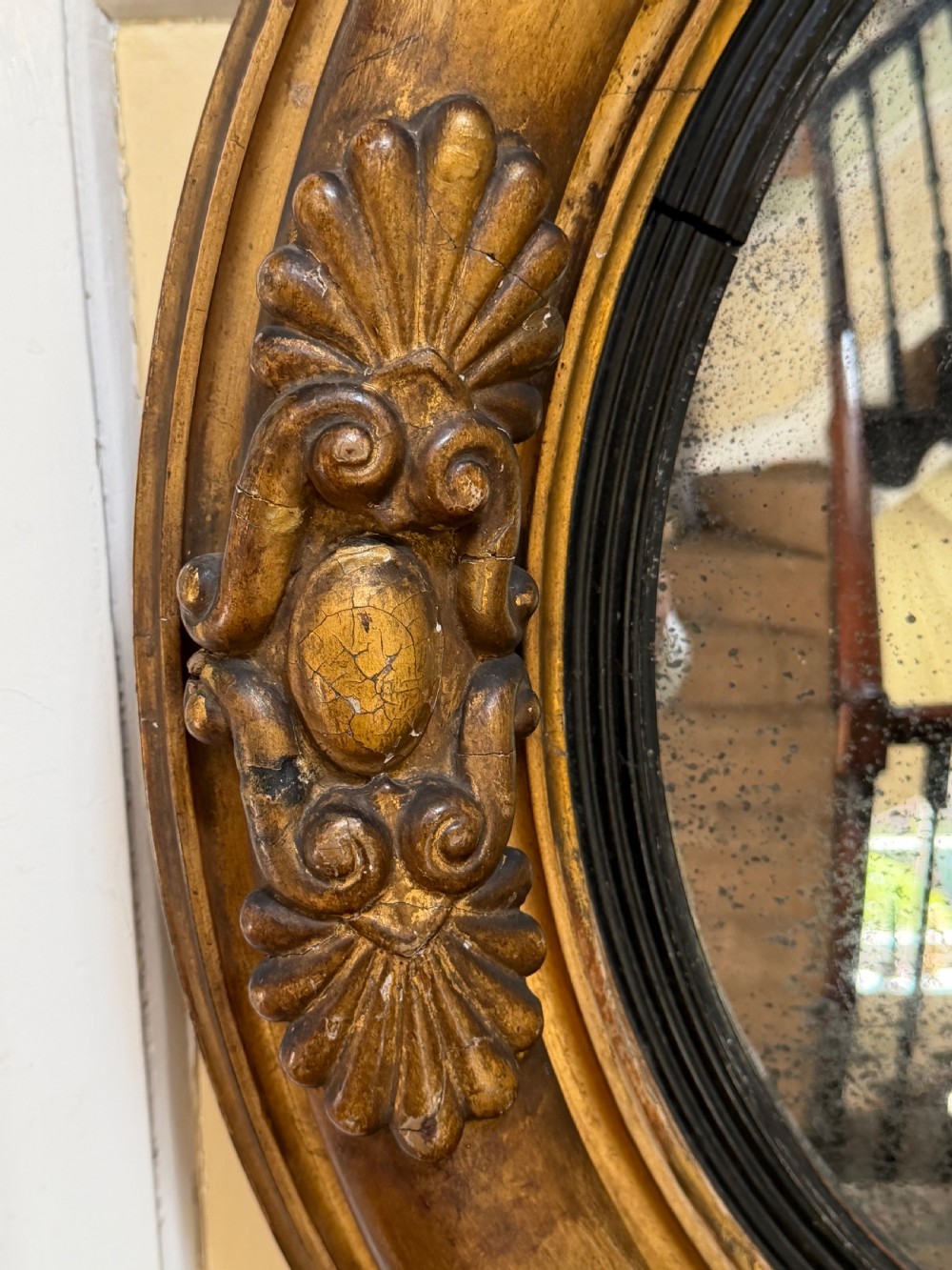 An early 19th-century Regency circular giltwood convex wall mirror featuring carved shell motifs, an ebonised reeded inner slip, and foxed mirror glass. View XI