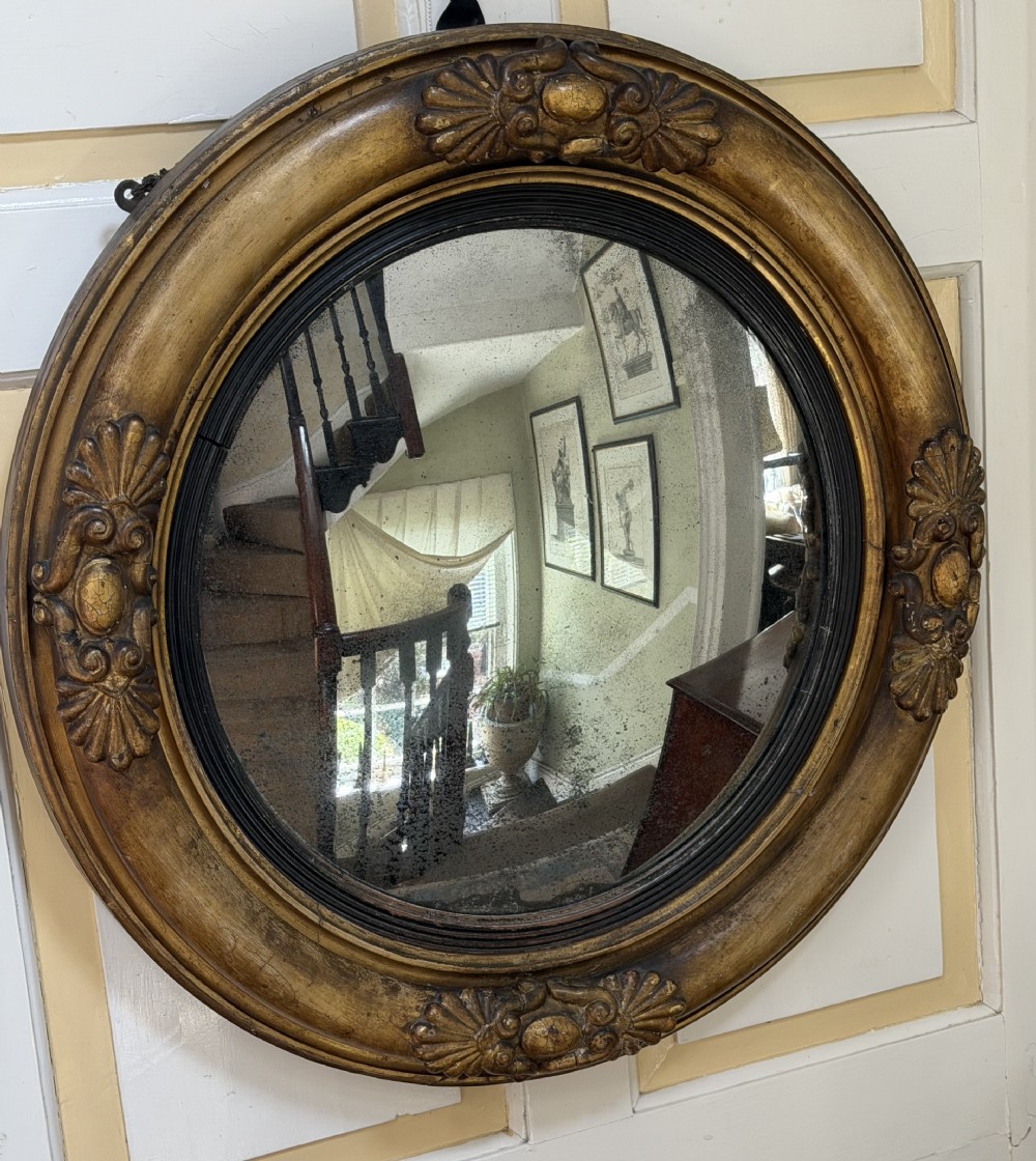 An early 19th-century Regency circular giltwood convex wall mirror featuring carved shell motifs, an ebonised reeded inner slip, and foxed mirror glass. View III