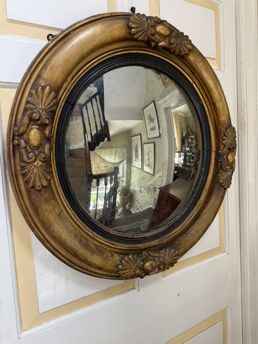An early 19th-century Regency circular giltwood convex wall mirror featuring carved shell motifs, an ebonised reeded inner slip, and foxed mirror glass. View XIII