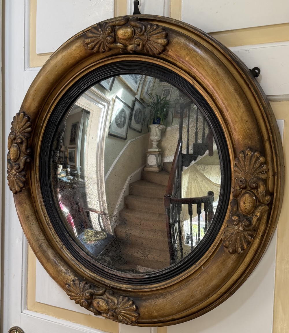 An early 19th-century Regency circular giltwood convex wall mirror featuring carved shell motifs, an ebonised reeded inner slip, and foxed mirror glass. View X