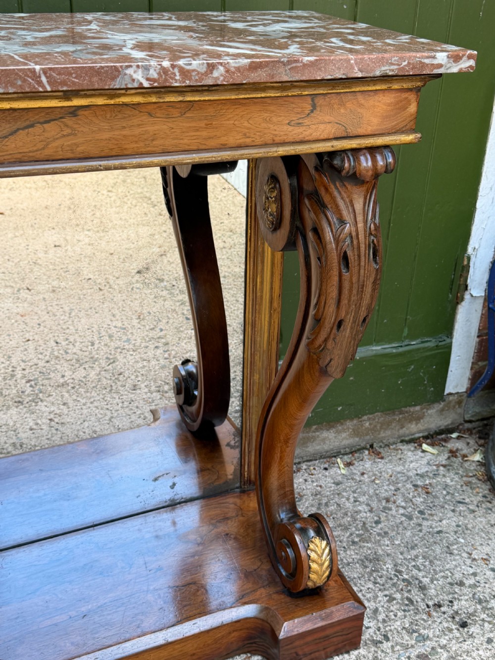 A Regency rosewood console table featuring a figured marble top, carved scroll legs with parcel-gilt details, an inset mirror back, and a plinth base. View V