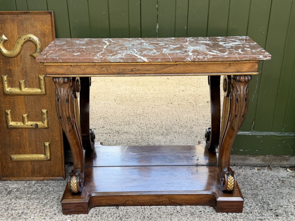 A Regency rosewood console table featuring a figured marble top, carved scroll legs with parcel-gilt details, an inset mirror back, and a plinth base. View XXVI