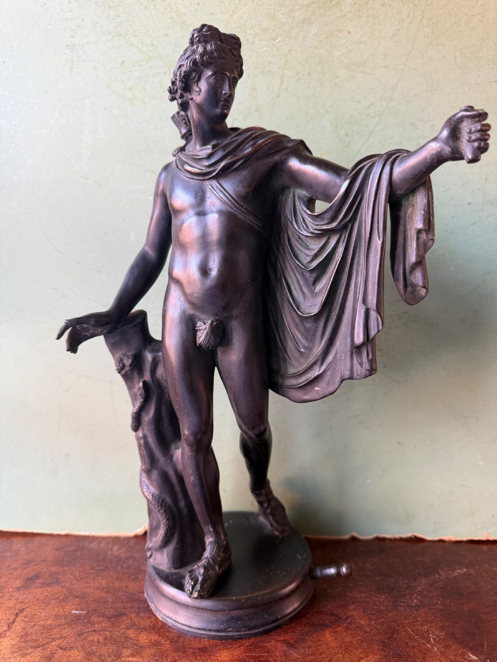 A dark patinated bronze figure depicting the classical god Apollo standing nude with drapery over his arm on a round base.
