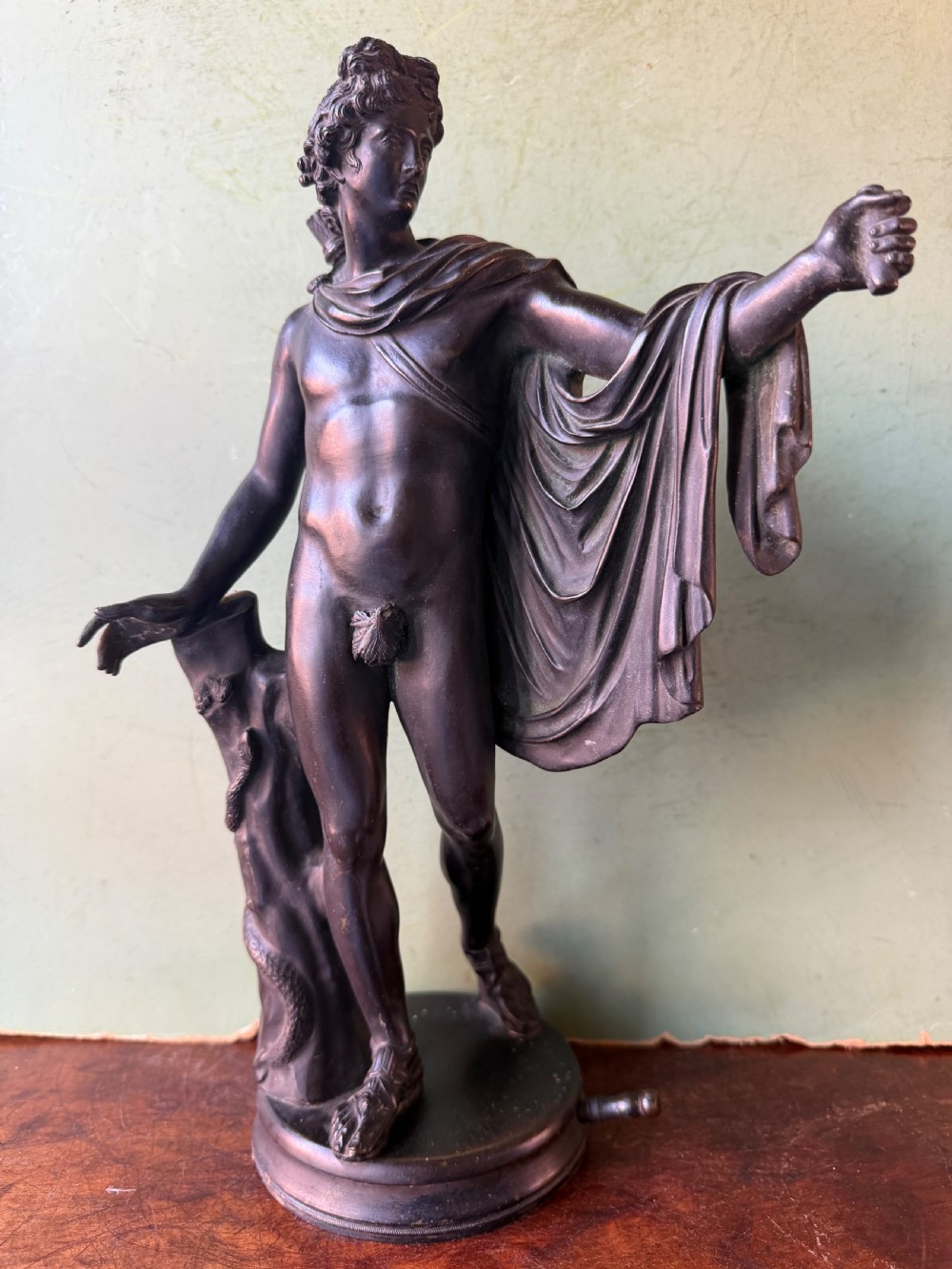 A dark patinated bronze figure depicting the classical god Apollo standing nude with drapery over his arm on a round base. View II