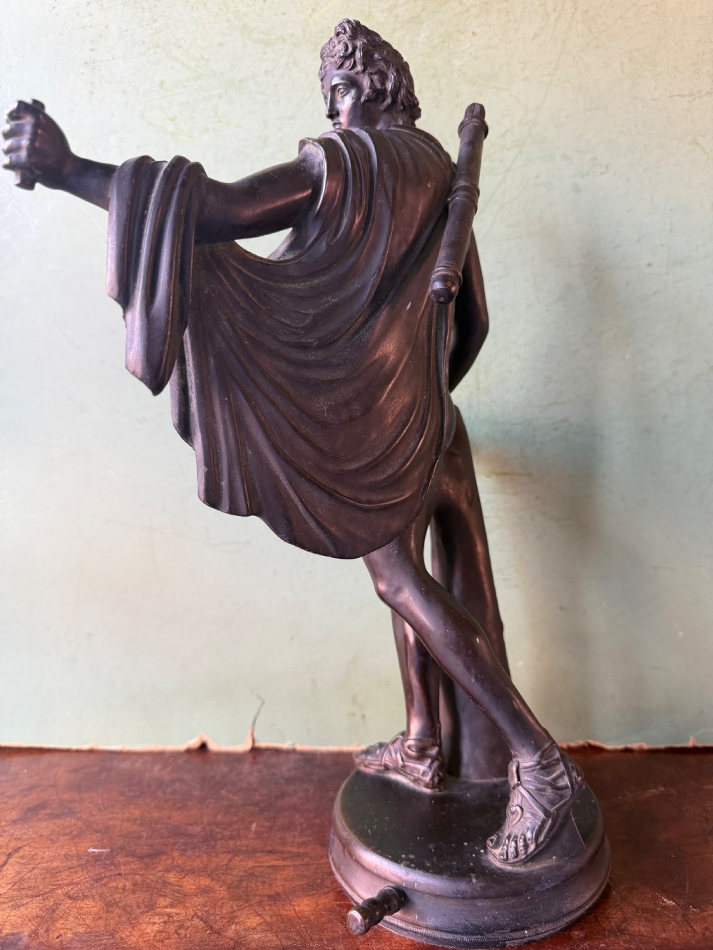 A dark patinated bronze figure depicting the classical god Apollo standing nude with drapery over his arm on a round base. View III