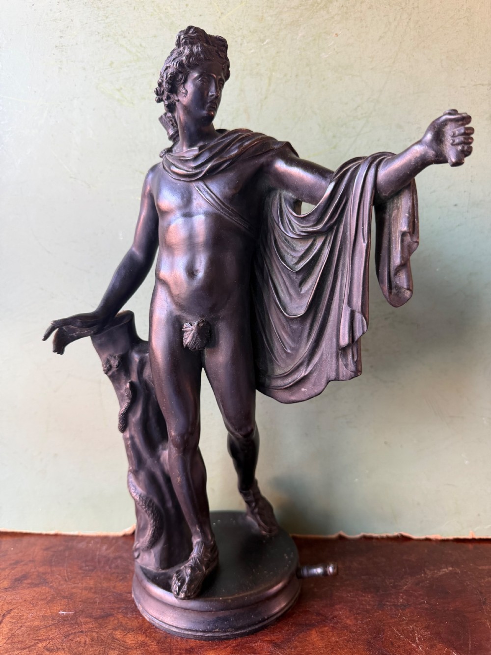 A dark patinated bronze figure depicting the classical god Apollo standing nude with drapery over his arm on a round base. View IV