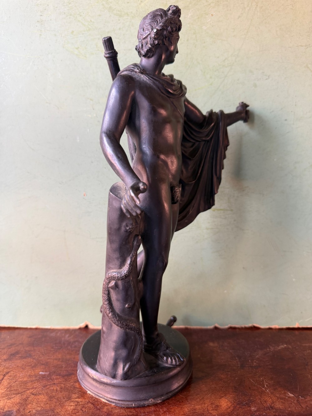 A dark patinated bronze figure depicting the classical god Apollo standing nude with drapery over his arm on a round base. View VI