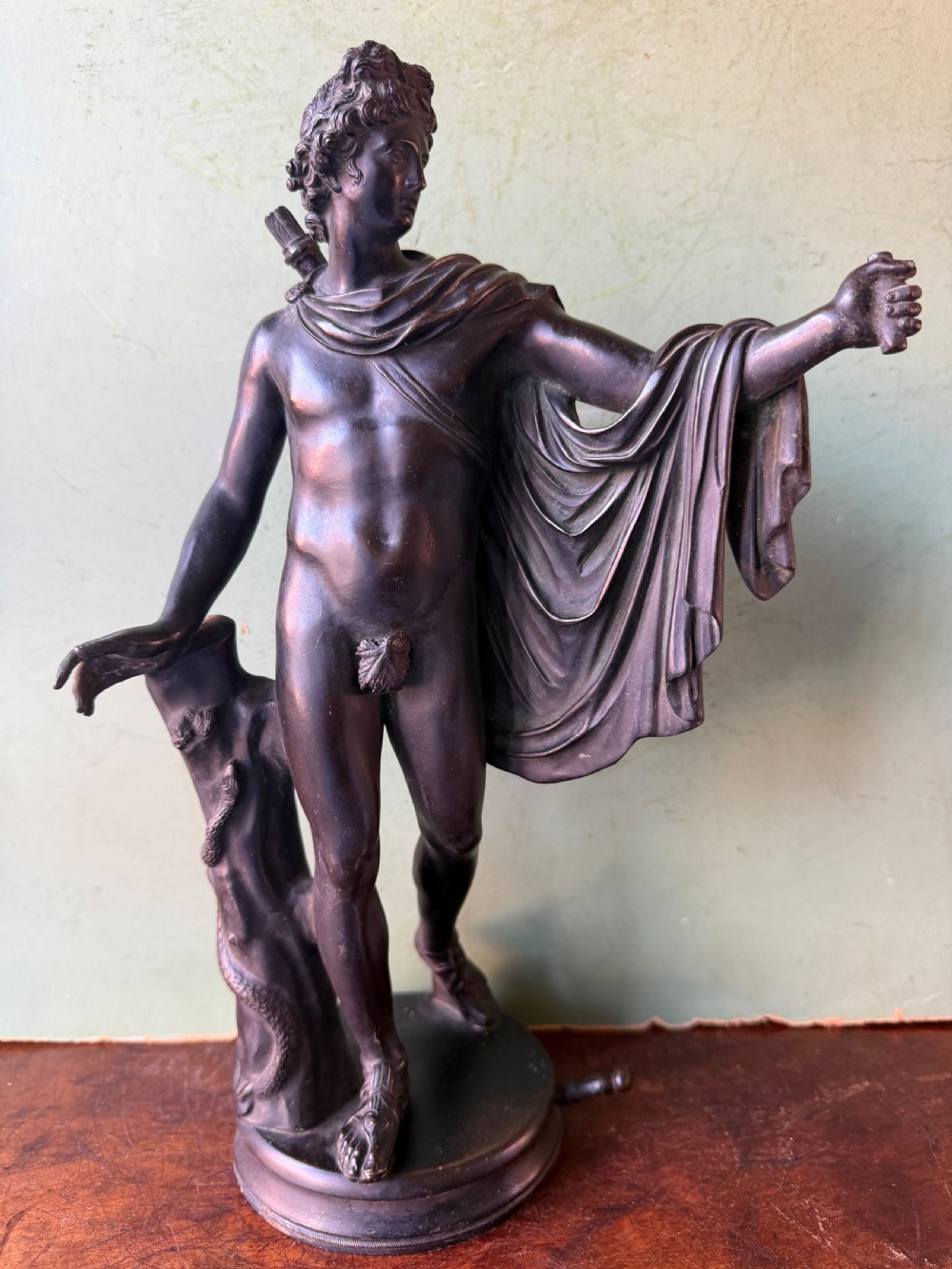 A dark patinated bronze figure depicting the classical god Apollo standing nude with drapery over his arm on a round base. View XV