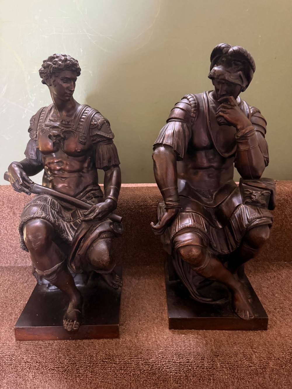 A pair of patinated dark bronze sculptures depicting two seated male figures in classical Roman armour mounted on rectangular bases.