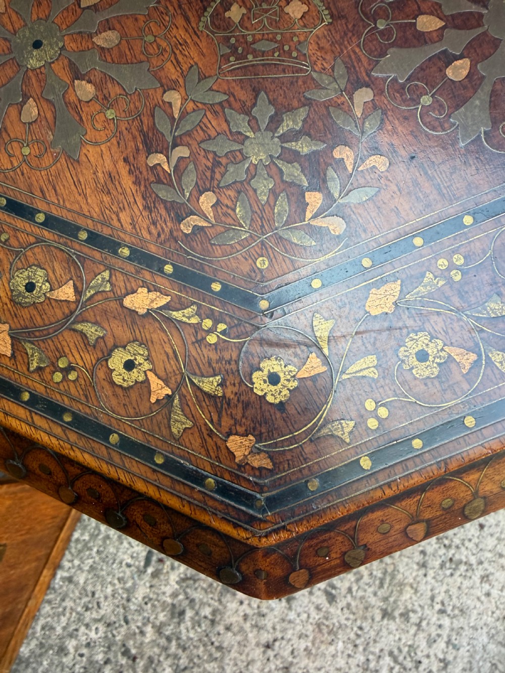 An octagonal Anglo-Indian teak side table featuring intricate floral and geometric brass and copper wire inlay on its top surface, set upon a carved and pierced folding lattice wooden base. View III