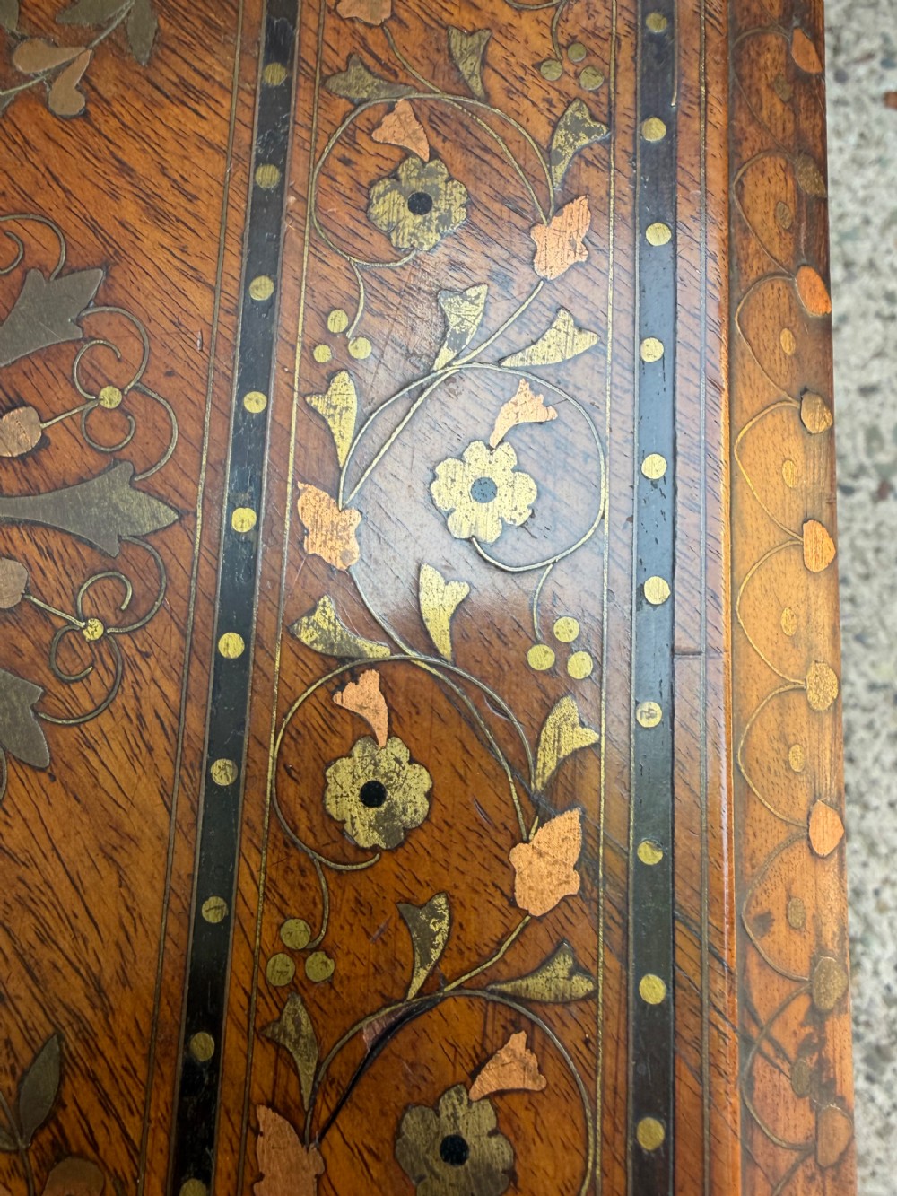 An octagonal Anglo-Indian teak side table featuring intricate floral and geometric brass and copper wire inlay on its top surface, set upon a carved and pierced folding lattice wooden base. View V