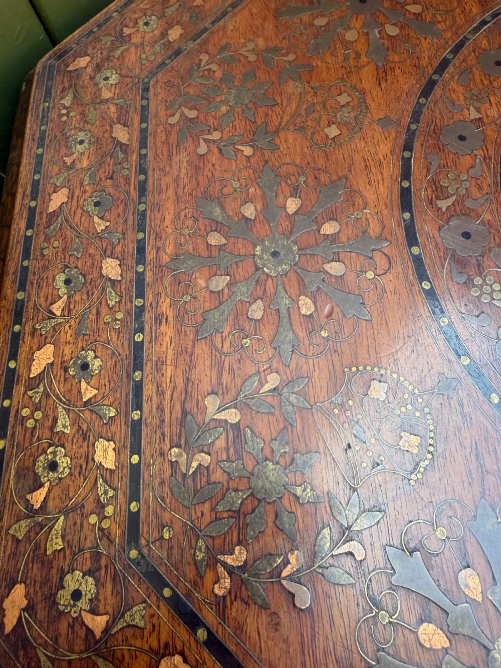 An octagonal Anglo-Indian teak side table featuring intricate floral and geometric brass and copper wire inlay on its top surface, set upon a carved and pierced folding lattice wooden base. View VI