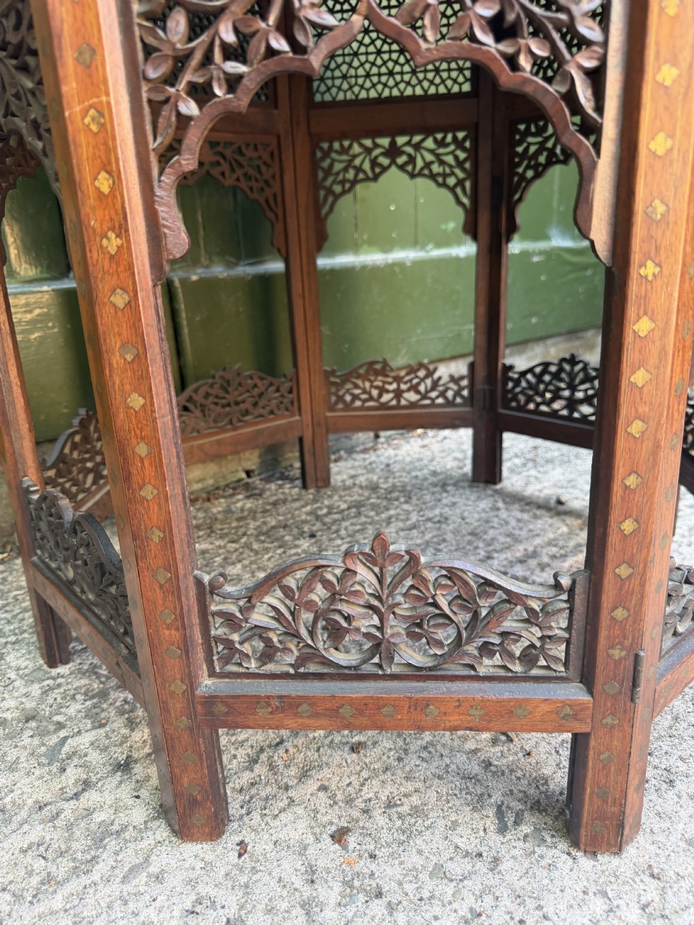 An octagonal Anglo-Indian teak side table featuring intricate floral and geometric brass and copper wire inlay on its top surface, set upon a carved and pierced folding lattice wooden base. View IX