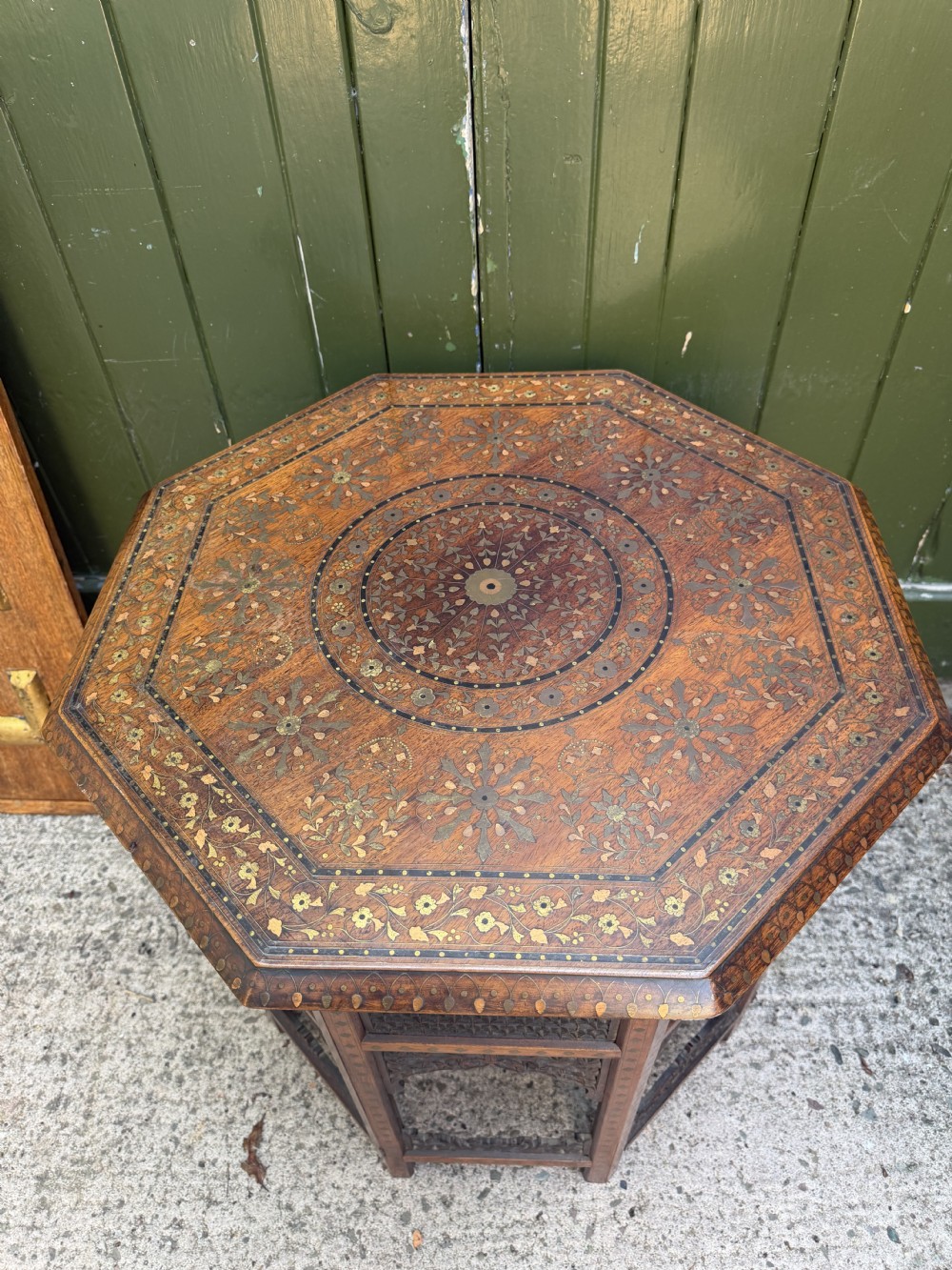 An octagonal Anglo-Indian teak side table featuring intricate floral and geometric brass and copper wire inlay on its top surface, set upon a carved and pierced folding lattice wooden base. View X