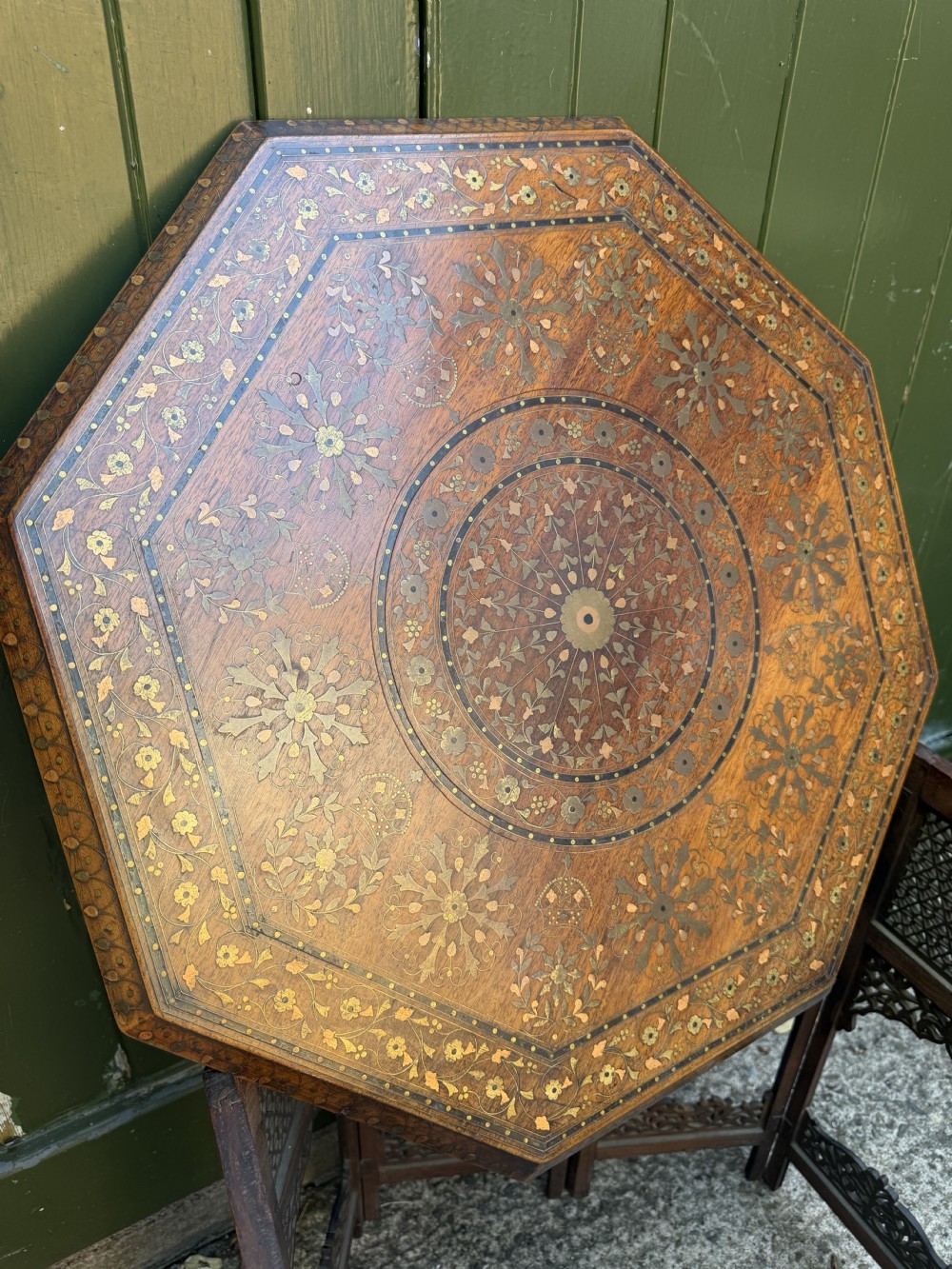 An octagonal Anglo-Indian teak side table featuring intricate floral and geometric brass and copper wire inlay on its top surface, set upon a carved and pierced folding lattice wooden base. View XI