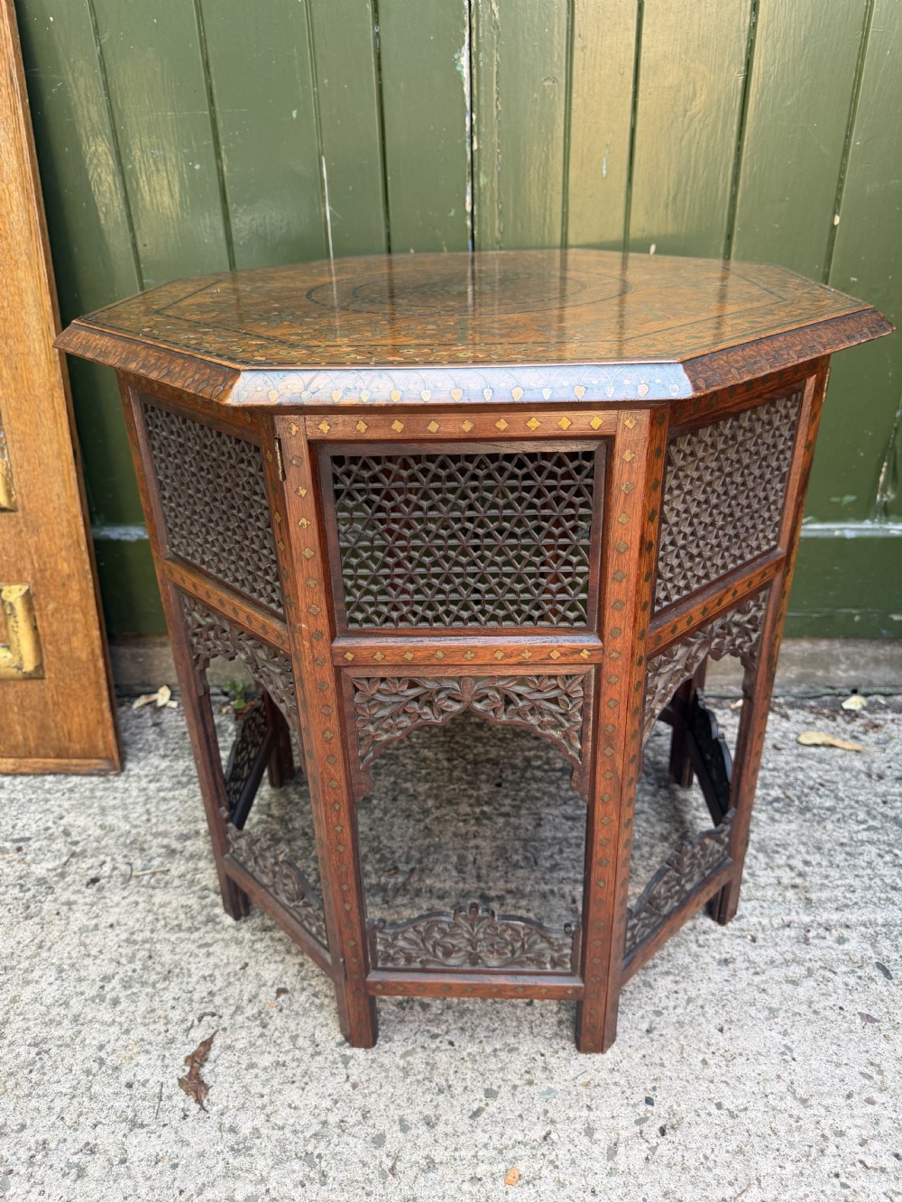 An octagonal Anglo-Indian teak side table featuring intricate floral and geometric brass and copper wire inlay on its top surface, set upon a carved and pierced folding lattice wooden base. View XVI
