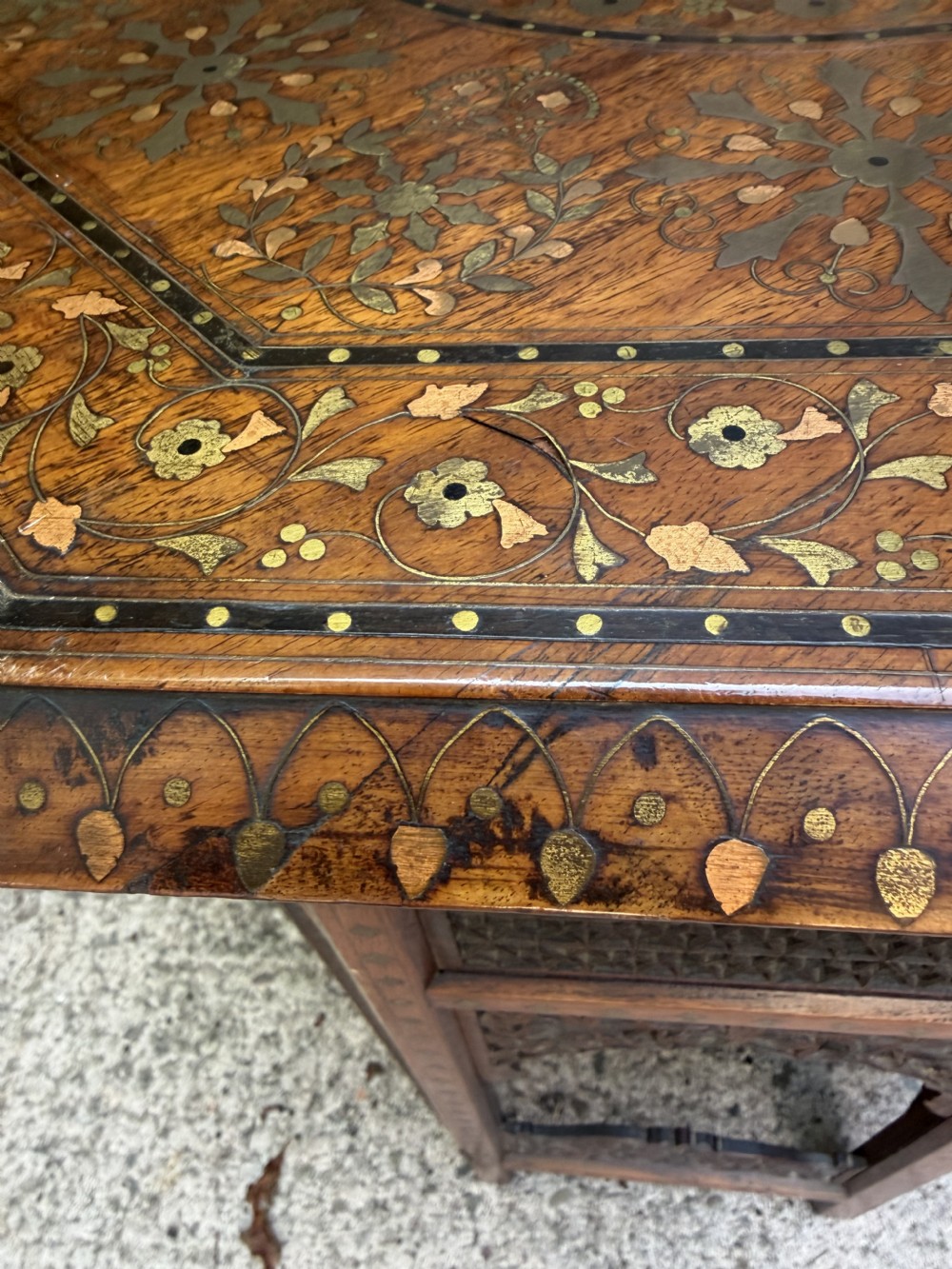 An octagonal Anglo-Indian teak side table featuring intricate floral and geometric brass and copper wire inlay on its top surface, set upon a carved and pierced folding lattice wooden base. View XVII