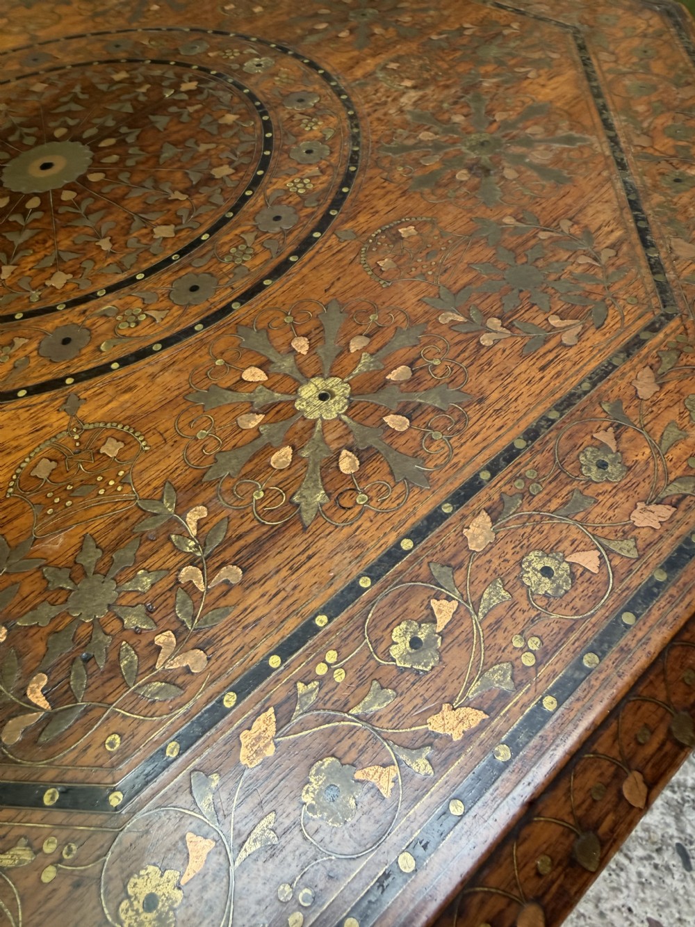 An octagonal Anglo-Indian teak side table featuring intricate floral and geometric brass and copper wire inlay on its top surface, set upon a carved and pierced folding lattice wooden base. View XVIII